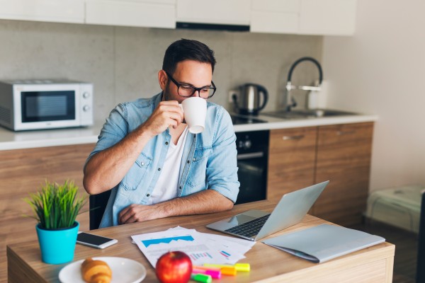 Man sitting in kitchen drinking coffee working on laptop